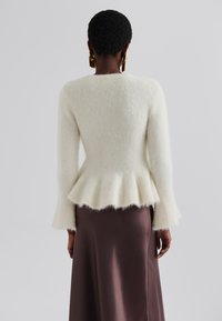 Pull blanc et moelleux, ajusté avec un ourlet en peplum, manches longues et finition texturée ; associé à une jupe en satin marron.