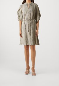 Helles graues Kleid mit strukturiertem Muster, kurzen Glockenärmeln, bindbarem Ausschnitt und verstellbarem Taillenbund. Kombiniert mit Riemchenabsätzen.