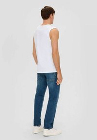 Young man facing away, wearing a white sleeveless shirt, blue jeans, and white sneakers on a plain light background.