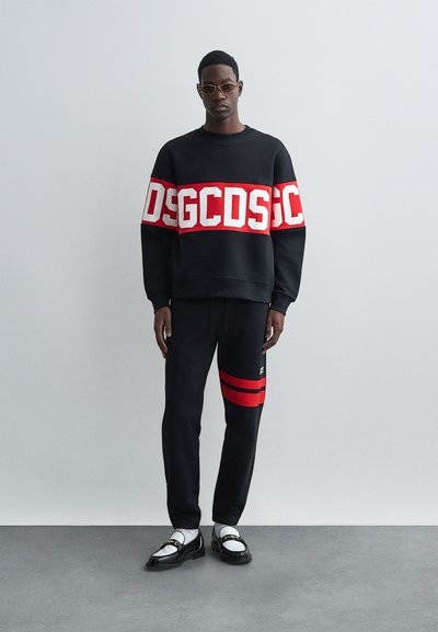 Μαύρη μπλούζα φούτερ με έντονα κόκκινα γράμματα μεγάλης γραμματοσειράς, σε συνδυασμό με μαύρες φόρμες που διαθέτουν κόκκινες ρίγες. Γυαλιστερά μαύρα παπούτσια και λευκές κάλτσες.