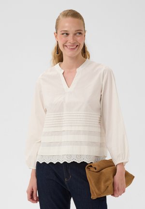 Femme souriante portant une blouse blanche à manches longues avec des plis et un ourlet en dentelle, jean foncé, tenant une pochette en daim marron pliée.