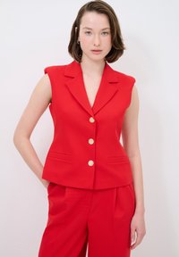 Roter ärmelloser Blazer mit Reverskragen, ausgestattet mit goldenen Knöpfen und seitlichen Taschen, kombiniert mit passenden roten Hosen. Glatte Textur.