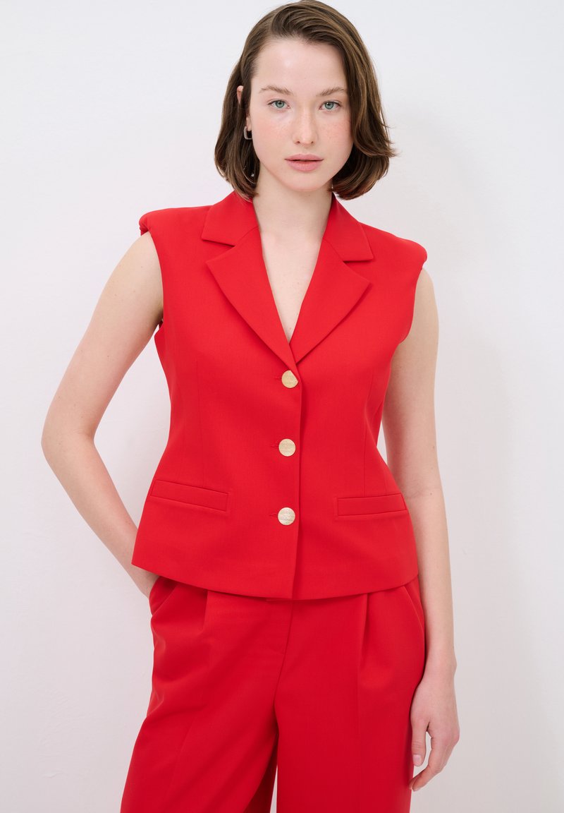 Roter ärmelloser Blazer mit Reverskragen, ausgestattet mit goldenen Knöpfen und seitlichen Taschen, kombiniert mit passenden roten Hosen. Glatte Textur.