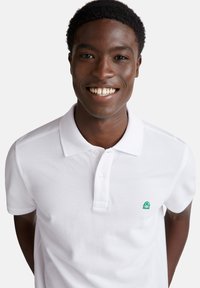 Camisa polo blanca de tela texturizada, con un logo verde en el lado izquierdo del pecho y un cuello clásico con dos botones.