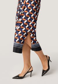 Patroon jurk met een geometrisch ontwerp in blauw, wit en oranje, met een split en een zwarte zoom. Gecombineerd met zwarte puntige hakken.