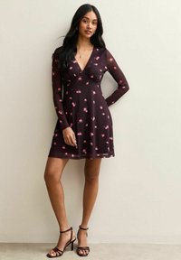 Vestito nero con un motivo floreale, maniche lunghe e trasparenti, scollatura a V, corpetto aderente e gonna svasata. Indossato con sandali con tacco a strappo.