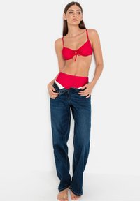 Red bikini set with a tie-front top, high-waisted bottom; paired with dark blue denim jeans. Smooth textures, casual fit, bare feet.