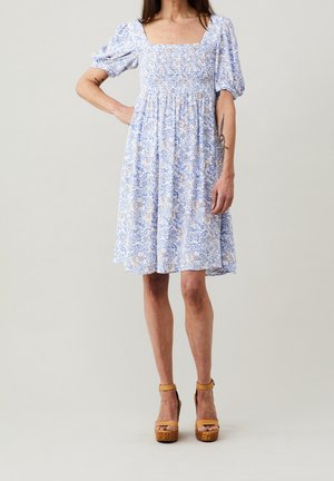 Kobieta ubrana w białą sukienkę do kolan z niebieskimi i beżowymi kwiatowymi wzorami, z bufkami na krótkich rękawach, kwadratowym dekoltem oraz beżowymi sandałami na platformie.