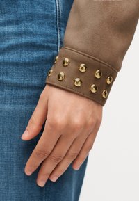 Χέρι που ακουμπάει σε μπλε τζιν, φορώντας μανίκι καφέ σουέντ μπουφάν με δύο σειρές λαμπερών χρυσών στρογγυλών τρουκς στη χειρολαβή.