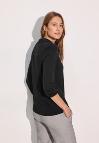 Schwarzes Langarmoberteil mit lockerer Passform, ausgestattet mit einem Kragen und umgeschlagenen Ärmeln, kombiniert mit grauen Jogginghosen. Glatte Textur, schlichtes Design.