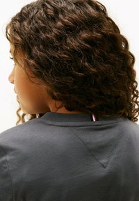 Eine Person mit lockigem Haar, seitlich und von hinten gesehen, die ein dunkelgraues Shirt mit einem kleinen rot-weißen und schwarzen Streifen am Kragen trägt.