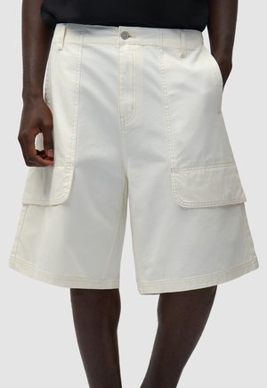 Uomo che indossa pantaloncini cargo bianchi fino al ginocchio con grandi tasche laterali e una maglietta nera, in piedi con le mani rilassate.