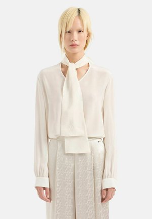 Blusa di seta beige chiaro con maniche lunghe e dettaglio con fiocco al collo, abbinata a pantaloni a vita alta con motivo coordinato.