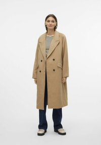 Cappotto doppiopetto color cammello con ampi rever, tasche laterali e bottoni neri, indossato sopra una camicia a righe e jeans.