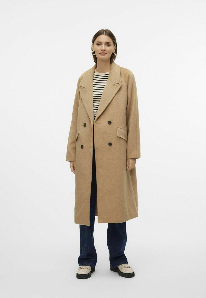 Manteau camel croisé avec grands revers, poches latérales et boutons noirs, superposé à une chemise rayée et un jean.