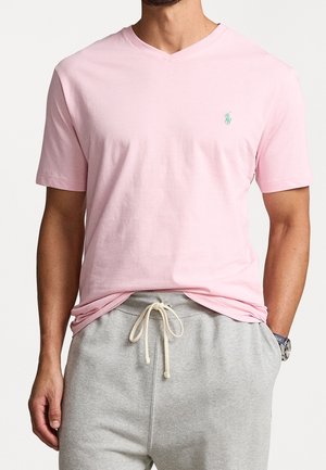 Rosa Baumwoll-T-Shirt mit V-Ausschnitt, ausgestattet mit einem kleinen grünen Logo auf der Brust, kombiniert mit grauen Sweatpants. Weicher Stoff und lässige Passform.