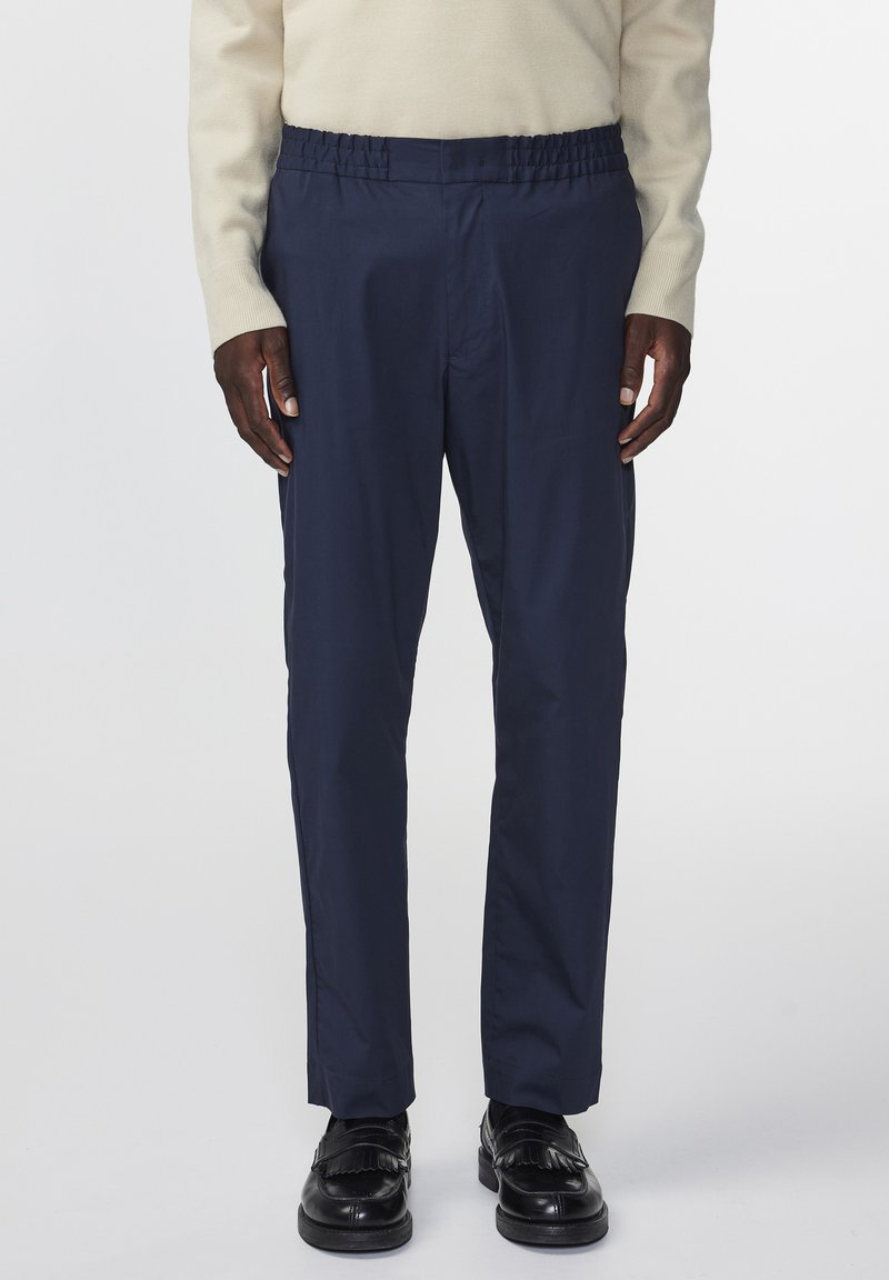 Marineblauwe broek met een elastische tailleband, rechte snit en een gladde textuur, gecombineerd met zwarte schoenen met een franjesdetail.
