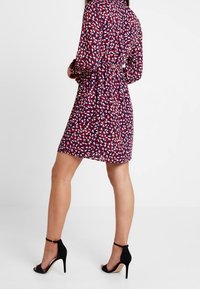 Kortärmad klänning med krage i marinblått, prydd med ett färgglatt lövmönster. Åtsmitande midja med en svävande kjol; matchad med svarta högklackade sandaler.