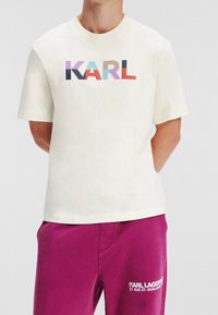 Cream cotton t-shirt featuring "KARL" in multicolored block letters. The model pairs it with dark magenta pants with a text logo.
