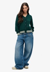 Zielony sweter z kapturem, z dekoltem w serek i sześcioma guzikami, z białymi paskami na dolnej krawędzi. Luźne niebieskie dżinsy o wyblakłym wykończeniu.