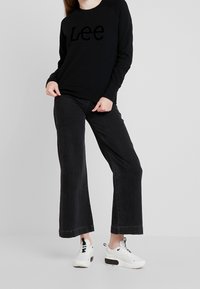 Persona vistiendo un sudadero negro de Lee, jeans negros de pierna ancha y zapatillas blancas, de pie contra un fondo gris claro liso.