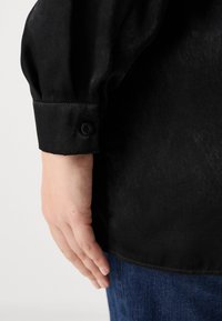 Black shirt with a smooth texture, featuring a buttoned cuff and a slightly loose fit. Paired with blue denim jeans.