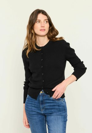 Woman with light brown wavy hair wearing a black buttoned cardigan and blue jeans, looking slightly to her left with hand in pocket.
