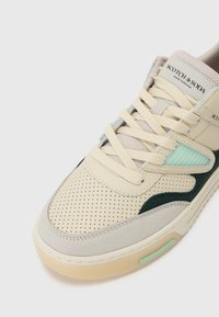 Beige sneaker with perforated upper, green and blue accents, flat laces, and textured details; logo on tongue; rubber sole with mint tag.