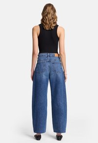 Pantalones de mezclilla de pierna ancha en azul medio con cintura alta, que cuentan con dos bolsillos traseros y un bajo sin rematar. Combinados con una camiseta negra ajustada.