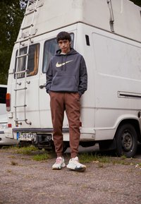 Harma Nike-huppari, jossa on iso logo, ruskeat verkkohousut ja moniväriset Nike-lenkkarit, seisoo valkoisen pakettiauton vieressä ulkona.