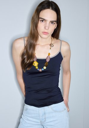 Jonge vrouw met lang bruin haar draagt een zwart tanktopje, lichtblauwe spijkerbroek en een kleurrijke kralenhalsketting, staand tegen een effen muur.