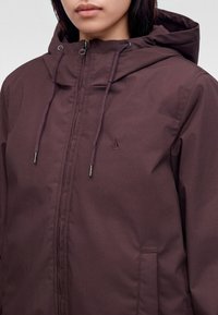 Femme portant une veste à capuche violet foncé avec une fermeture éclair et des cordons, visible du cou jusqu'au milieu du torse.