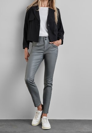 Femme debout contre un mur gris, portant une veste courte noire, une chemise blanche, un pantalon gris ajusté et des baskets blanches.