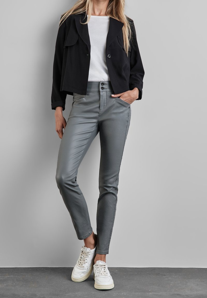 Femme debout contre un mur gris, portant une veste courte noire, une chemise blanche, un pantalon gris ajusté et des baskets blanches.