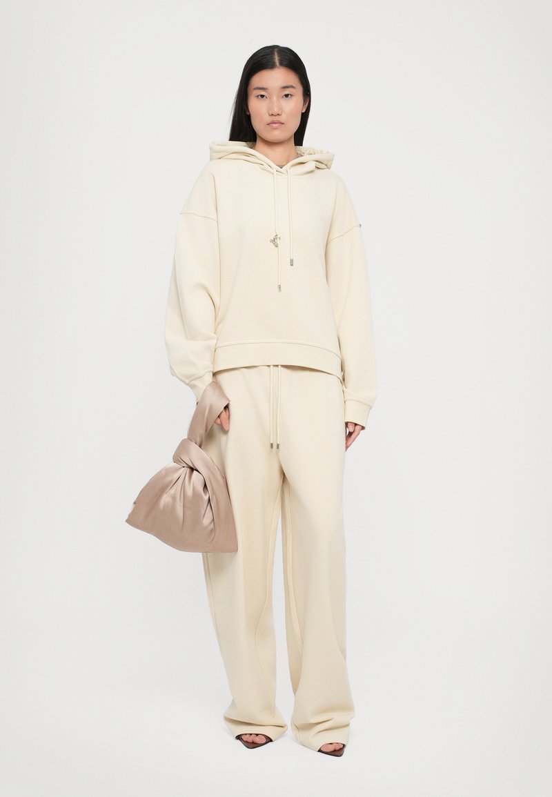 Beige oversized hoodie with front pocket and adjustable drawstring, paired with matching tapered sweatpants. Holding a taupe satin pouch bag.