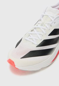 White athletic shoe with black accents and mesh upper, featuring a textured surface and a bright pink sole component. Laces are prominent.