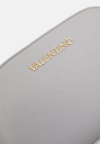 Borsa cosmetica in pelle grigia chiaro con superficie texturizzata, con logo "VALENTINO" impresso in oro. Forma arrotondata, design minimalista.