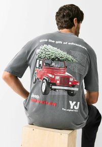 Szürke rövid ujjú póló, melyen egy piros Jeep Wrangler grafikája látható, tetején karácsonyfa, szöveggel kísérve.