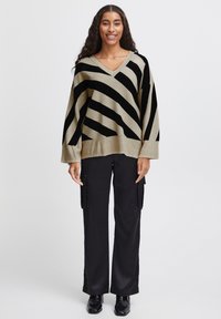 Pull tricot beige et noir à rayures avec un col en V, associé à un pantalon cargo noir. Le pull présente de larges rayures diagonales et une coupe ample.
