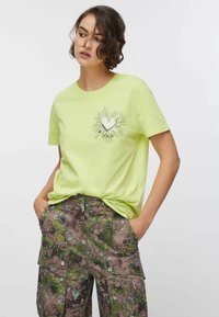 Limonkowa zielona koszulka z krótkimi rękawami i srebrnym sercem otoczonym promieniami słonecznymi na piersi. W zestawie spodnie cargo w kwiatowy wzór.