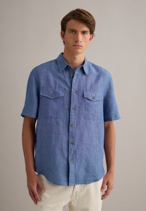 Junger Mann, der ein kurzärmliges blaues Hemd mit Brusttaschen trägt, vor einem einfarbigen beigen Hintergrund stehend.