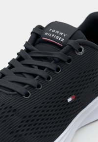 Sneaker en maille noire avec un design à lacets, dotée d'une languette grise portant le logo "TOMMY HILFIGER" et d'un petit détail du drapeau sur le côté. Semelle blanche.