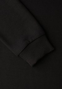 Schwarzer, gerippter Strickstoff zeigt einen Teil eines Ärmels und der Manschette. Die Textur ist fein mit vertikalen Linien und sitzt eng am Handgelenk.