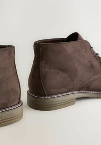 Botas de ante marrón hasta el tobillo con punta redonda y detalles cosidos, colocadas una al lado de la otra sobre un fondo claro.