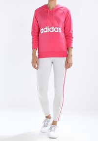 Rosa huvtröja med vit "adidas"-logotyp, komplett med grå leggings med rosa sidostreck. Vita sneakers fullföljer outfiten.