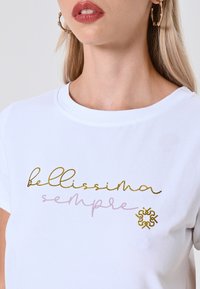 T-shirt bianca in cotone con la scritta "bellissima sempre" in testo corsivo oro e rosa, e un emblema floreale decorativo dorato sul lato.