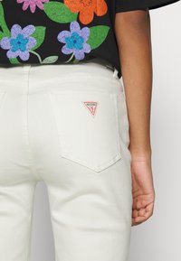 Pantalones de denim blanco con bolsillos traseros y un parche de logo en forma de triángulo rojo. La tela tiene una textura suave y un diseño minimalista.