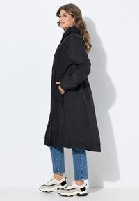 Svart vattentät jacka med huva, hel längd, med dragkedja fram, sidofickor och lös passform, kombinerad med blå denim och kraftiga sneakers.