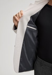 Blazer grigio chiaro con un'esterno testurizzato e fodera interna in satin scuro; presenta una piccola tasca interna e un design leggero.