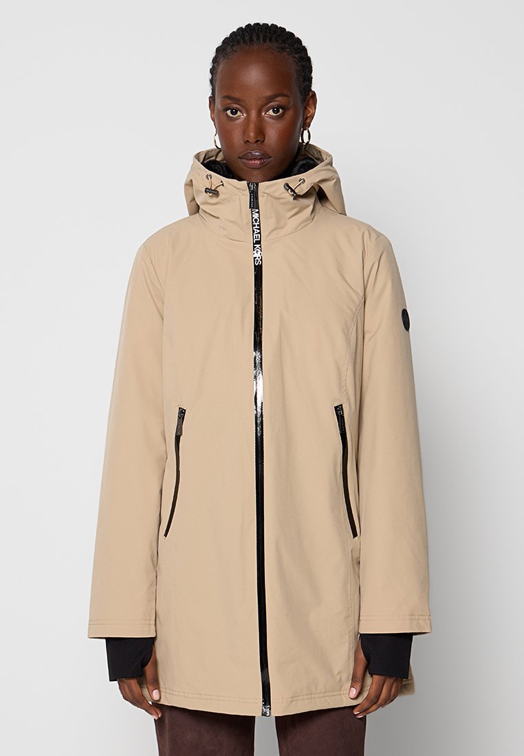 Beige waterdichte jas met een strakke capuchon, zwarte ritsdetails en zijzakken. Gladde textuur met een relaxte pasvorm.